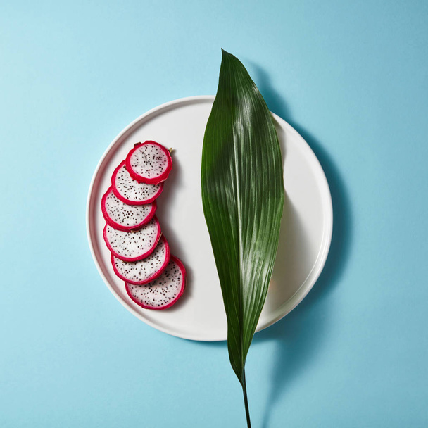 Several pieces of a sliced white pink pitahaya on white plate with green leaf on blue background. Flat lay and copy space - Fotoğraf, Görsel