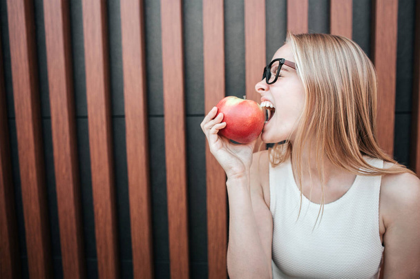 Kuva blondista tytöstä, joka puree palaa punaisesta omenasta. Hän nauttii hetkestä. eristetty raidallisella pohjalla
. - Valokuva, kuva