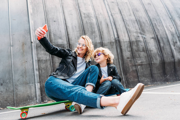 mother and son sitting on skateboards on street and taking selfie with smartphone - Foto, immagini