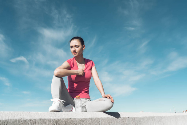 confident slender girl sitting on the ground in the sun, resting and gaining strength after training - Foto, Imagem