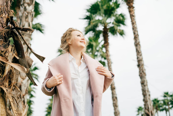 joyful young woman in a stylish pink coat walking in a park next to green palms - Foto, Imagen