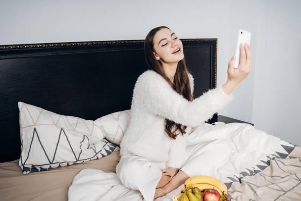beautiful long-haired girl in pajamas sitting on bed making selfie on her smartphone - Photo, image