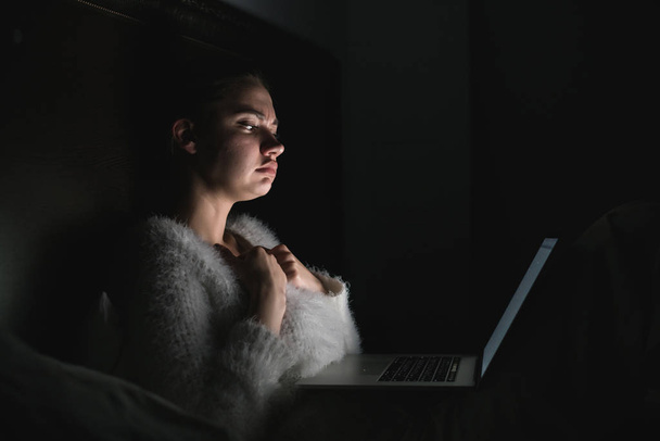 young sad girl sitting in bed late at night, watching a scary movie on her laptop - Фото, изображение