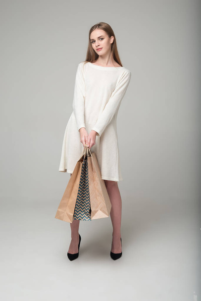 Jovem modelo de cabelos longos menina loira em branco vestido curto stands segurando pacotes de compras
 - Foto, Imagem