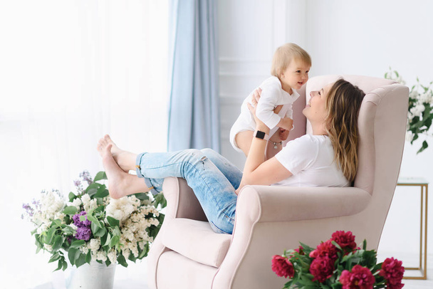 A young mother plays with her young child in a soft armchair in her large and bright room - Foto, immagini