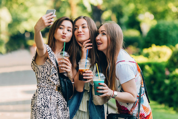 Happy friends in the park on a sunny day . Summer lifestyle portrait of three multiracial women enjoy nice day, holding glasses of milkshakes. Taking picture on mobile phone. Best friends girls having - Foto, imagen