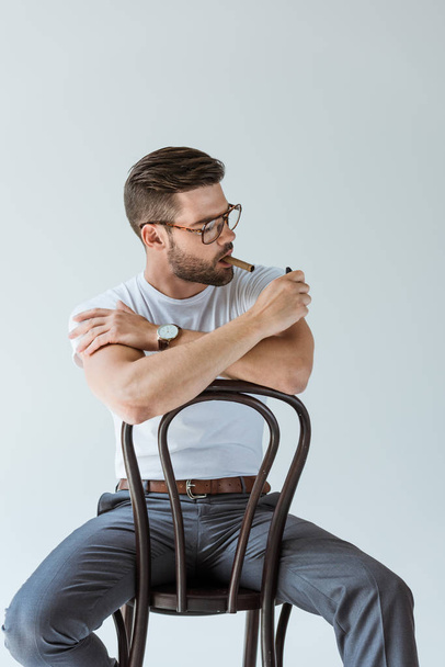 Szép szakállas férfi világítás szivart, miközben ül a székre, elszigetelt fehér background - Fotó, kép