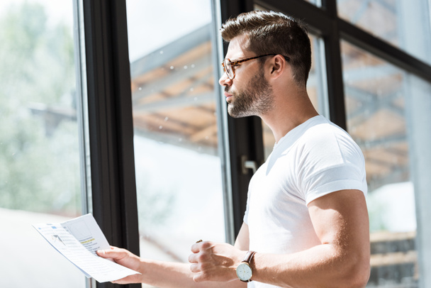 Handsome bearded businessman in glasses reading report by window - Foto, afbeelding