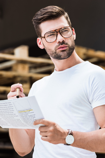 Fashionable confident businessman reading business document and holding cigar by wooden palettes - Photo, Image