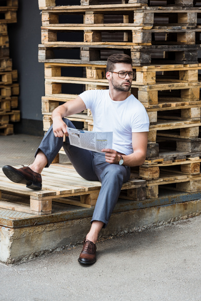 Handsome bearded businessman sitting on wooden palettes with cigar and report in hands - Photo, Image