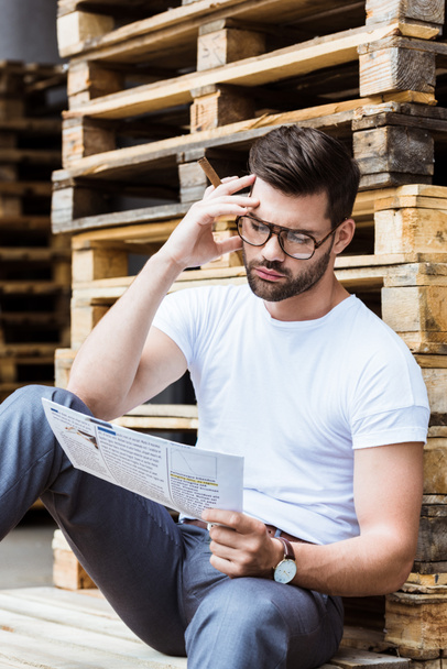 Homme d'affaires confiant à la mode dans des lunettes fumant le cigare tout en lisant le document d'affaires par des palettes en bois
 - Photo, image