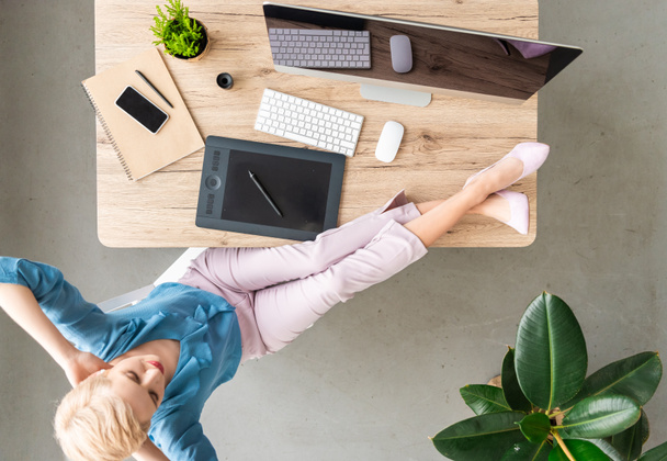вид сверху на стильную женщину-телеработницу, сидящую с ногами на столе с компьютером, смартфоном и графическим планшетом дома
  - Фото, изображение