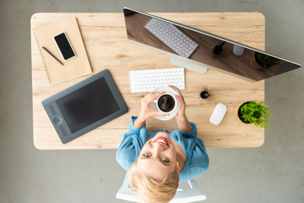 グラフィック タブレット、スマート フォン、自宅のコンピューターとオフィス カメラとテーブルでのコーヒーのホールディング カップを見て笑みを浮かべて女性フリーランサーの立面図  - 写真・画像