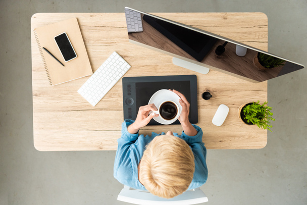 overhead view of female freelancer holding cup of coffee at table with graphic tablet, smartphone and computer at home office  - Foto, Imagem