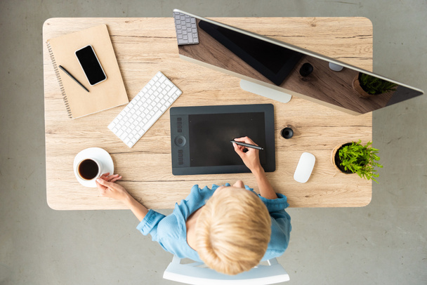 υπερυψωμένη θέαση της γυναικείας teleworker χρήση tablet γραφικών στο τραπέζι με το φλυτζάνι του καφέ, το smartphone και τον υπολογιστή στο γραφείο στο σπίτι  - Φωτογραφία, εικόνα