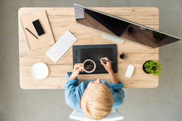 над головой вид женщины-фрилансера, пьющей кофе, работающей на графическом планшете за столом со смартфоном и компьютером в домашнем офисе
  - Фото, изображение