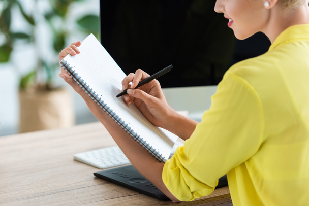 imagem recortada de uma mulher freelancer sorrindo escrevendo em livro didático à mesa com tablet gráfico e computador
 - Foto, Imagem