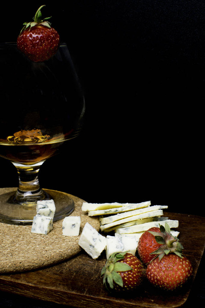Still life with Cognac and french blue cheese on a wooden board. Glass with cognac, cheese, strawberry on a wooden background - Photo, image