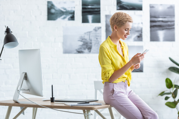 вид сбоку улыбающейся женщины-фрилансера, использующей смартфон рядом со столом с графическим планшетом и компьютером в домашнем офисе
  - Фото, изображение