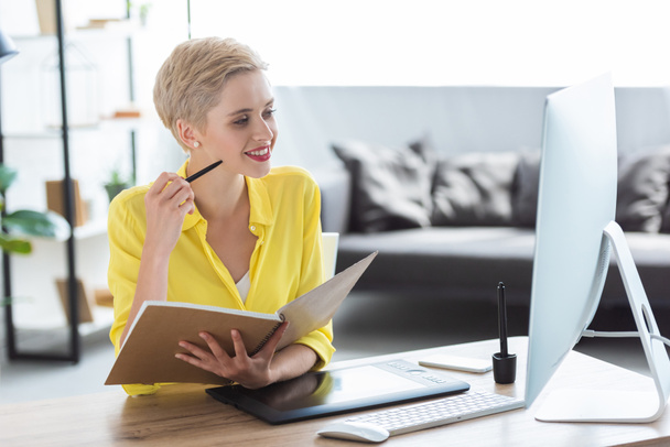 pensativo freelancer femenino sosteniendo libro de texto y sentado a la mesa con computadora y tableta gráfica
  - Foto, imagen