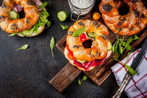 Variety of homemade bagels sandwiches with sesame and poppy seeds, cream cheese,  ham, radish, arugula, cherry tomatoes, cucumbers, with ingredients on dark concrete background copy space top view - Photo, Image