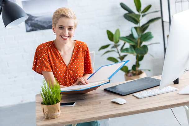female freelancer holding book at table with graphic tablet and computer in home office  - Фото, изображение