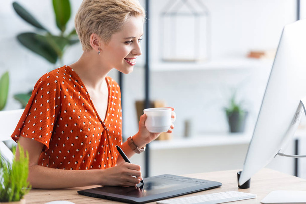 幸せな女性フリーランサー グラフィック タブレット コンピューターとテーブルで図面のコーヒー カップの側面図  - 写真・画像