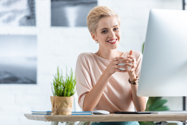 Улыбающаяся деловая женщина с чашкой кофе за столом с графическим планшетом и компьютером в офисе
  - Фото, изображение