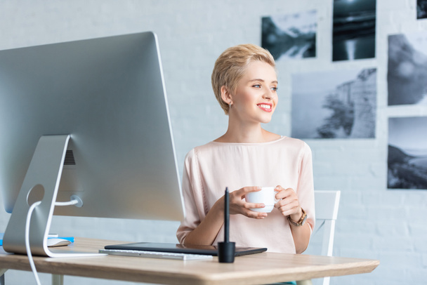 グラフィック タブレットとホーム オフィスのコンピューターとテーブルでのコーヒーの女性フリーランサー ホールディング カップを笑顔  - 写真・画像
