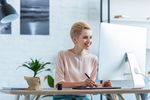sorridente freelance femminile che disegna su tavoletta grafica a tavola con computer
  - Foto, immagini