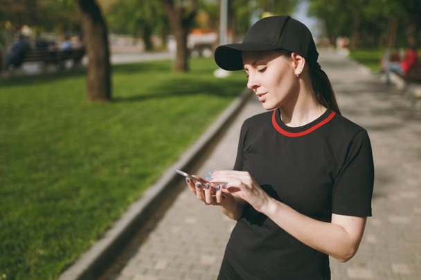 Şehir park açık havada yolda Smartphone, genç atletik güzel esmer kız siyah üniforma ve cep telefonuyla eğitim sırasında kap ayakta. Fitness, sağlıklı yaşam konsepti - Fotoğraf, Görsel