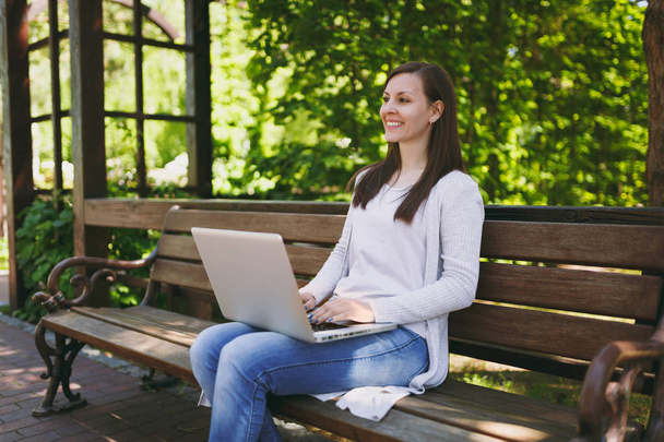 Young successful businesswoman in light casual clothes. Woman sitting on bench working on modern laptop pc computer in city park in outdoors on nature. Mobile Office. Freelance business concept - Fotó, kép