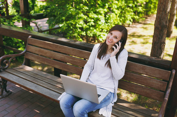 Young happy female in light casual clothes talking on mobile phone. Woman sitting on bench working on modern laptop pc computer in street outdoors on nature. Mobile Office. Freelance business concept - Foto, Bild
