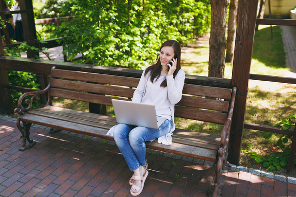 携帯電話で話しているカジュアルな軽装で若い幸せな女性。自然の屋外ストリートで現代のラップトップ pc に取り組んでのベンチに座っている女性。モバイル オフィス。フリーランスのビジネス コンセプト - 写真・画像