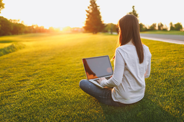 Молодая женщина вид сзади. Женщина работает за компьютером ноутбука с пустым чёрным экраном, чтобы скопировать пространство в городском парке на лужайке на зеленой траве. Мобильный офис. Концепция внештатного бизнеса
 - Фото, изображение