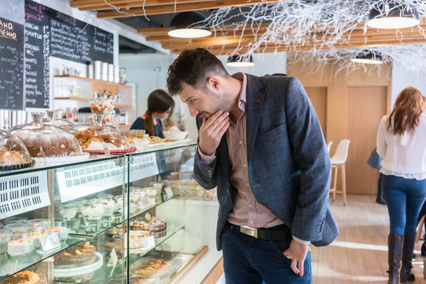Homem indeciso olhando para vários bolos expostos na vitrine
 - Foto, Imagem