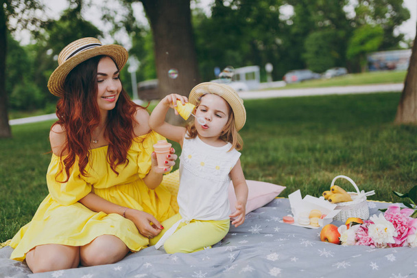 Joyful woman in yellow dress play on green grass in park, rest with little cute child baby girl hold soap bubble blower. Mother, little kid daughter. Mother's Day, love family, parenthood, childhood - Photo, Image