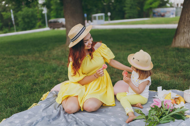 Smiling woman in yellow clothes rest and eat macaroons dessert with little child baby girl on green grass lawn in park. Mother, little kid daughter. Mother's Day, love family, parenthood, childhood - Fotoğraf, Görsel