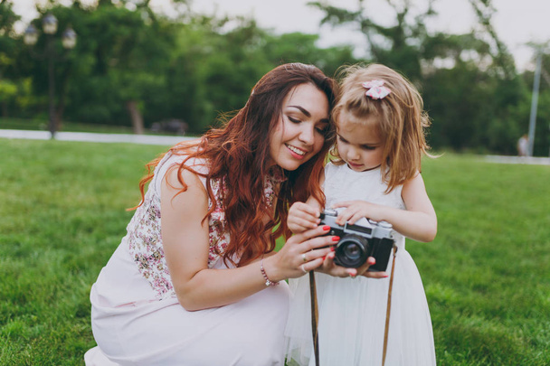 Smiling woman in light dress and little cute child baby girl holding retro vintage photo camera in green park. Mother, little kid daughter. Mother's Day, love family, parenthood, childhood concept - Photo, Image