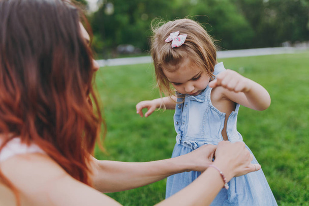 Woman with curly red hair dresses little pretty child baby girl in denim dress on green grass in city park. Mother, little kid daughter. Mother's Day, love family, parenthood, childhood concept - Foto, imagen