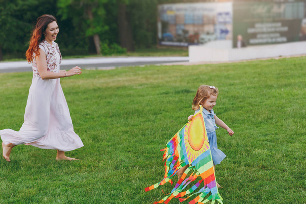Laughing woman in light dress and little cute child baby girl running, catching up and play with colorful kite in park. Mother, little kid daughter. Mother's Day, love family, parenthood, childhood - 写真・画像