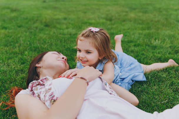 Smiling woman in light dress and little cute child baby girl lie on green grass lawn in park rest and have fun. Mother, little kid daughter. Mother's Day, love family, parenthood, childhood concept - Photo, Image