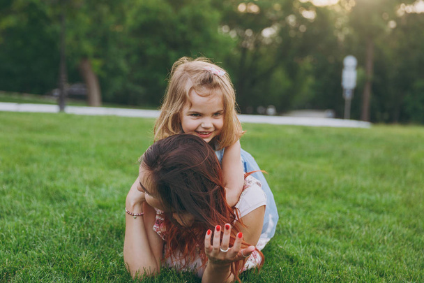 Woman in light dress and smiling little cute child baby girl lie on green grass in park rest play and have fun. Mother, little kid daughter. Mother's Day, love family, parenthood, childhood concept - Photo, Image