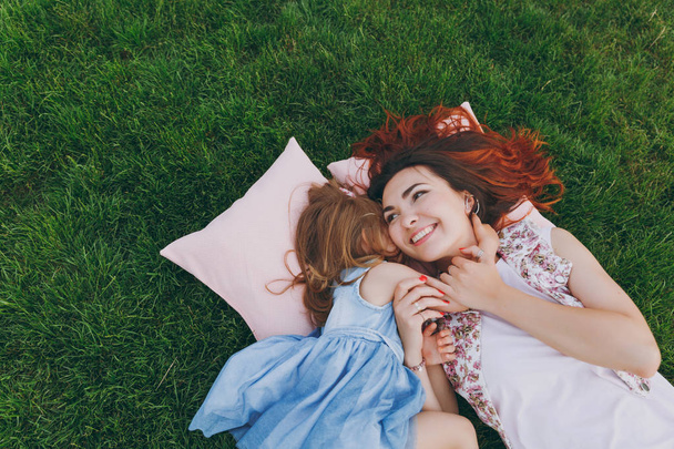 Smiling woman and little cute child baby girl lie on pillows on grass in park hug, embrace and rest, have fun. Mother, little kid daughter. Mother's Day, love family, parenthood, childhood concept - Photo, Image