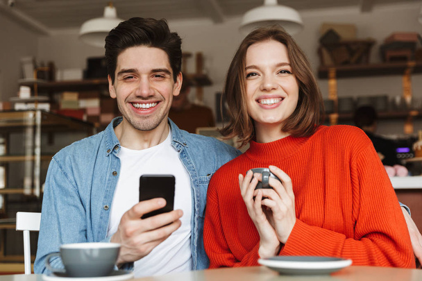 Smiling young couple sitting at the cafe table drinking coffee and holding mobile phone - Foto, Bild