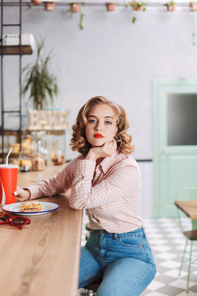 Young thoughtful lady in shirt and jeans sitting at the bar counter with coca cola glass and dremily looking in camera in cafe - Foto, afbeelding