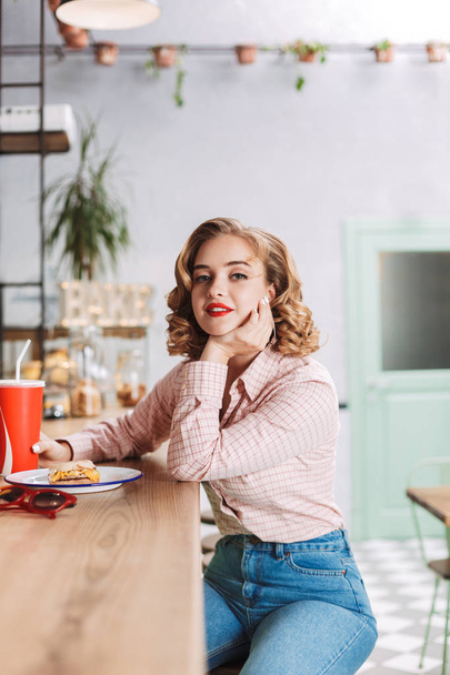 Beautiful lady in shirt and jeans sitting at the bar counter with coca cola glass and dremily looking in camera in cafe - Foto, imagen