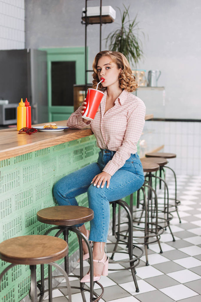 Belle jeune femme en chemise et jeans assis au comptoir du bar et boire de l'eau gazeuse tout en regardant dans la caméra dans le café
 - Photo, image