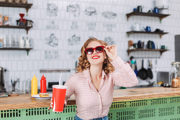 Portrait of smiling lady in sunglasses and shirt sitting at the bar counter with soda water in hand and happily looking in camera in cafe - Foto, imagen