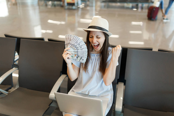 Περιχαρής ταξιδιώτη τουριστικά γυναίκα που εργάζεται σε φορητό υπολογιστή λαβή δέσμη δολάρια μετρητά χρήματα κάνουν νικητής χειρονομία περιμένετε στο λόμπι hall στο αεροδρόμιο. Επιβάτες που ταξιδεύουν στο εξωτερικό για απόδραση το Σαββατοκύριακο. Air πτήση έννοια - Φωτογραφία, εικόνα